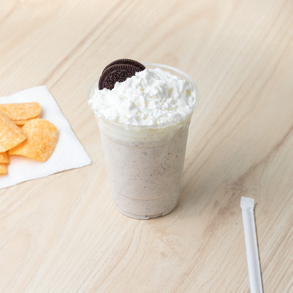A 16 oz UNIQ clear PET plastic cup with a cookies and cream milkshake and whipped cream on a wooden table next to a snack, demonstrating versatile beverage and food pairing for wholesale cafes and bistros.